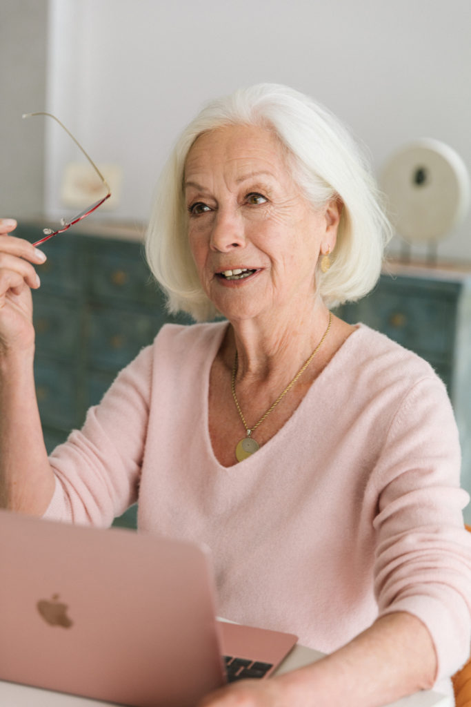 Scilla holds her glasses while looking up from her computer as if talking to someone near by.